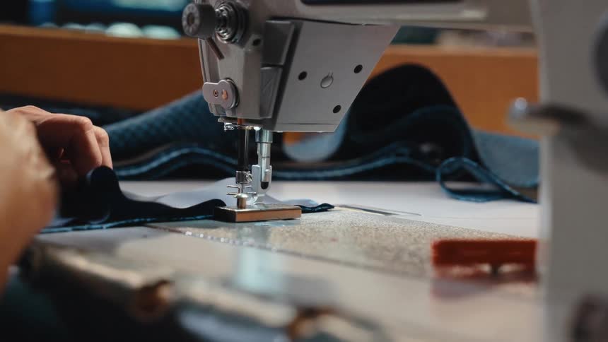 An over-the-shoulder view shows a person carefully sewing a piece of blue fabric on a professional machine