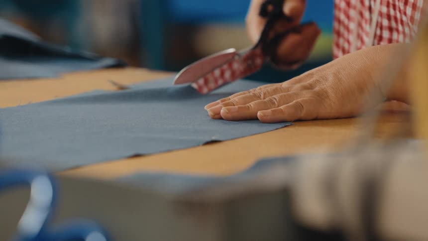 Slow motion close-up of a garment worker's hands using large shears to cut a pattern from blue fabric on a workshop table