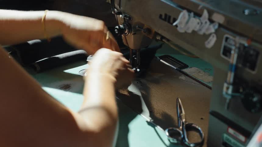 A tailor's hands guide dark fabric through an industrial sewing machine in a sunlit workshop