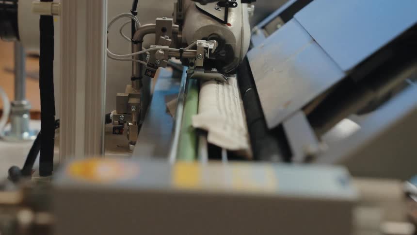 A close-up, low-angle shot of an industrial sewing machine automatically stitching a piece of fabric in a factory setting