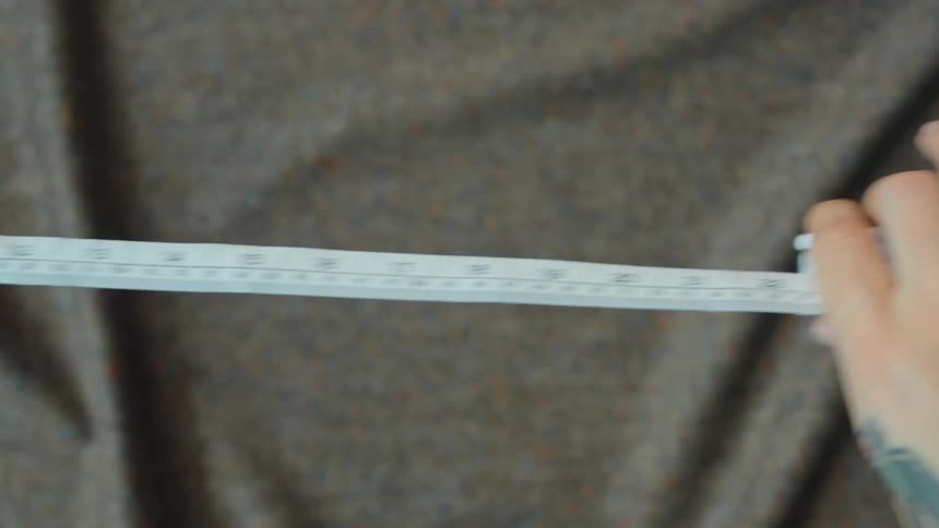 A close-up of a tattooed hand extending and retracting a white tailor's tape measure against a fabric background