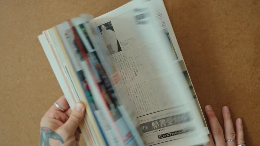A person with tattooed hands quickly flips through the pages of a vintage Japanese men's fashion magazine