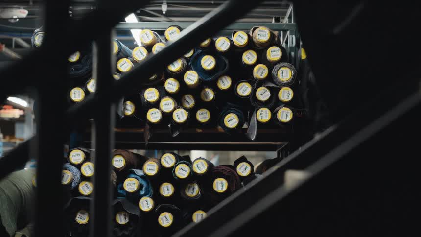 Labeled rolls of denim fabric are stacked on industrial shelves in a textile factory warehouse