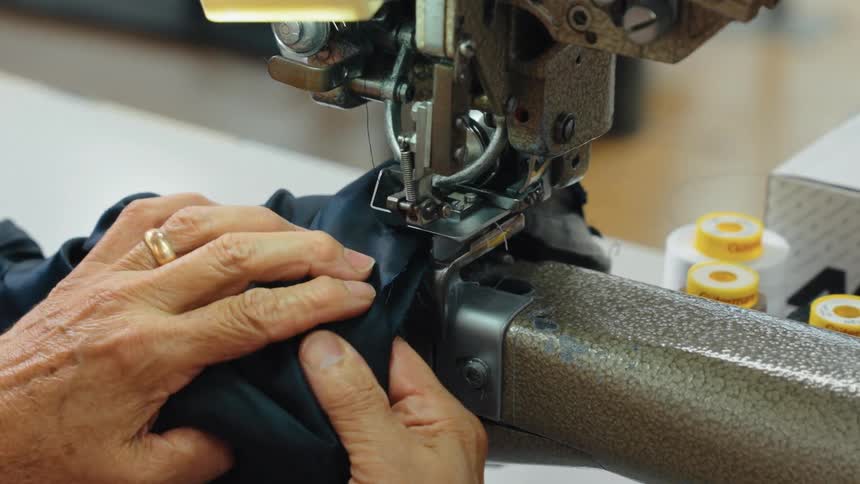 An experienced tailor carefully guides dark blue fabric through an industrial sewing machine in slow motion