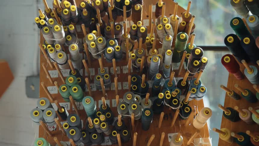 A wooden rack in a workshop is filled with dozens of organized spools of thread in various colors