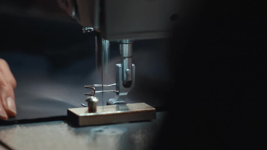 A detailed closeup shot of a sewing machine needle moving rapidly as a seamstress guides dark fabric through it