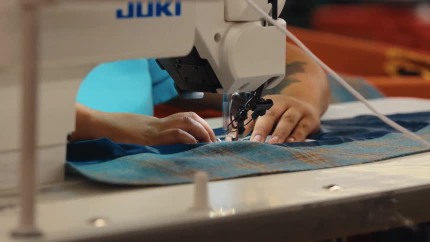 A close-up shot of a seamstress's hands with tattoos guiding blue plaid fabric through an industrial sewing machine in a workshop