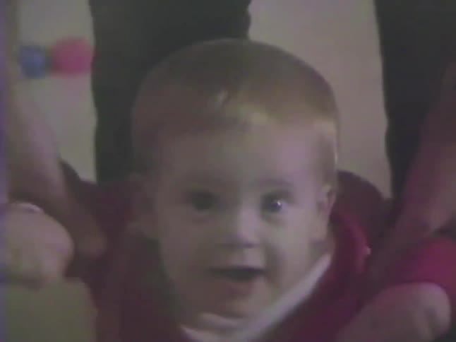 Vintage home video captures a close-up of a smiling baby being held up by their parent while learning to walk