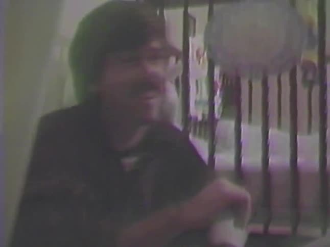A cute baby lies on the floor before their father helps them with their socks in a grainy home video from 1986