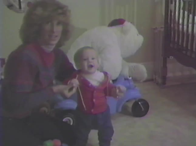 A mother helps her smiling baby learn to walk in their toy-filled room in a heartwarming vintage home movie