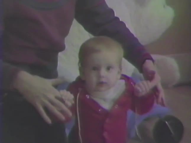 An adorable baby in a red sweater takes their first steps with help from a parent in a nostalgic home video