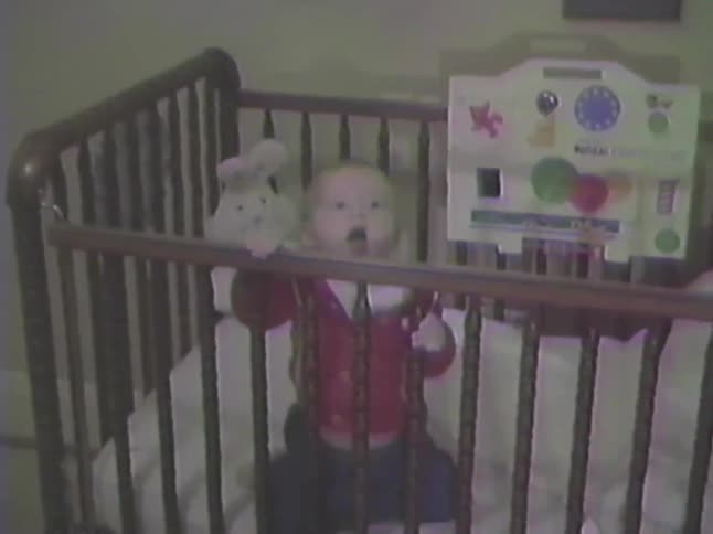 Vintage home video footage shows a cute baby in a red sweater standing up and yawning in a wooden crib