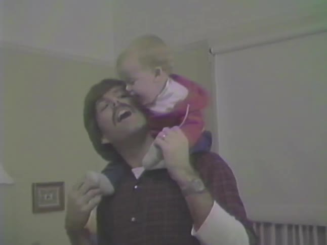 A joyful baby laughs while getting a shoulder ride from their dad who then playfully flips them upside down in a vintage home video