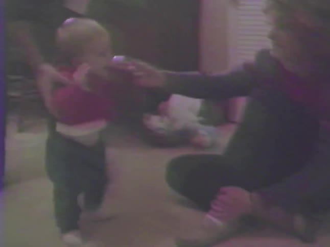 A sweet home video captures a baby's joyful reaction as a woman offers a toy while another adult helps the child stand