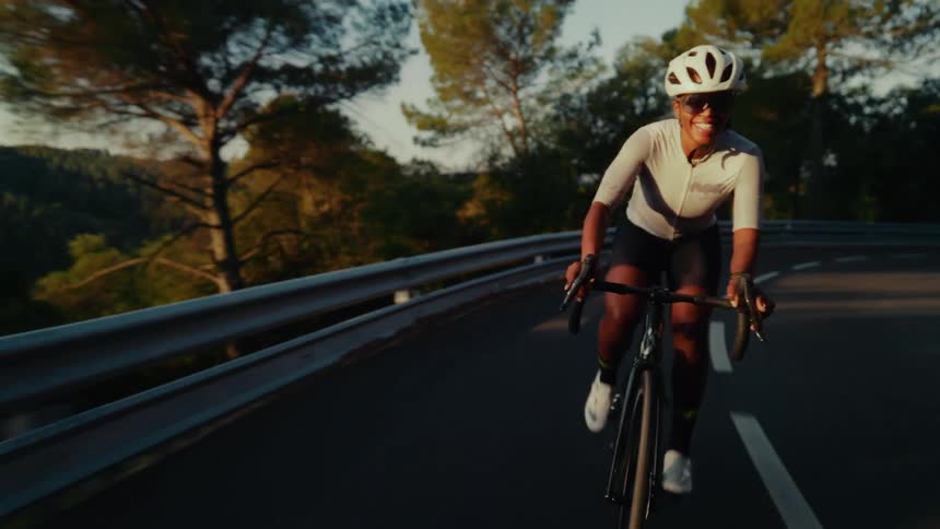 A smiling female road cyclist rides her bike down a winding mountain road during a beautiful golden hour sunset