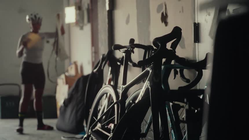 Bicycles are silhouetted in the foreground of a garage as a female cyclist prepares her gear in the background