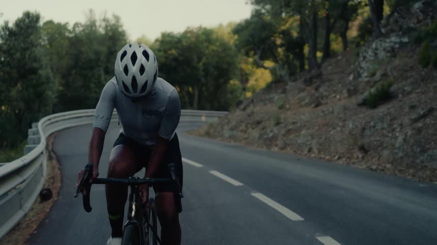 An athletic woman wearing a helmet and sunglasses laughs with joy and determination while cycling up a winding mountain road