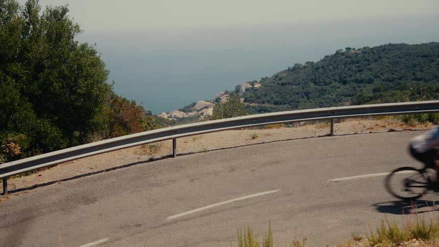 A road cyclist descends a winding mountain pass with a beautiful view of the sea on a sunny day