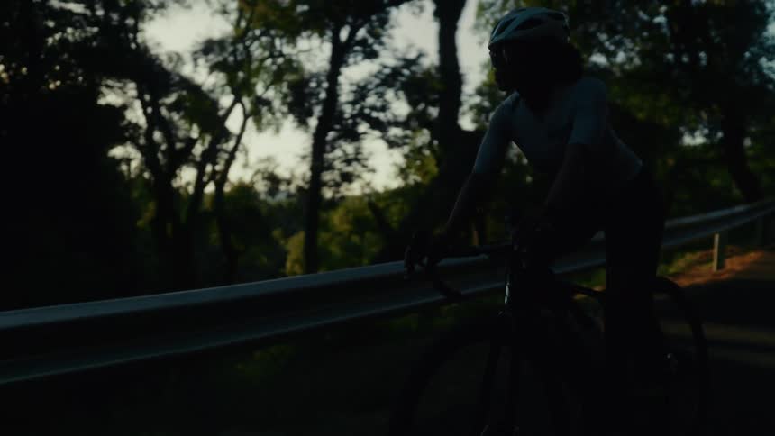 A determined female cyclist in a helmet rides her bicycle down a scenic road, silhouetted by the setting sun in slow motion