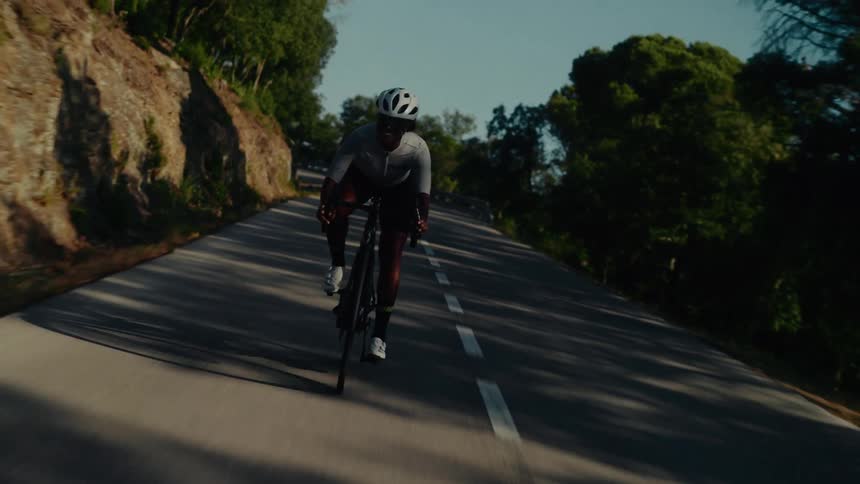 An athletic cyclist wearing a helmet and sunglasses rides a road bike up a winding country road in slow motion