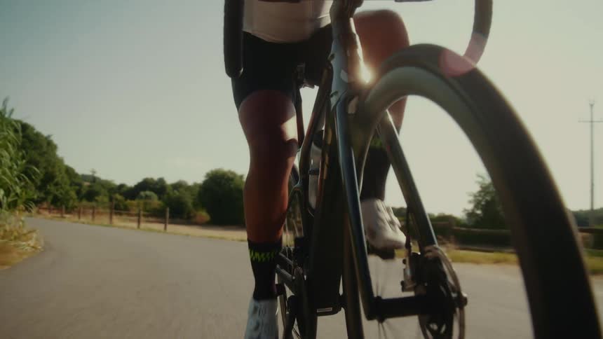 A dynamic low-angle close-up follows a cyclist's legs as they pedal a road bike along a paved country road in the bright sun