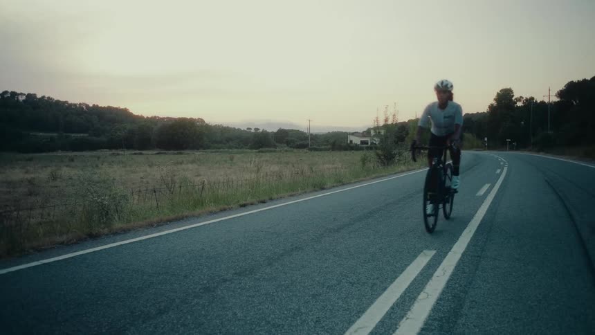 A determined female cyclist rides her road bike past the camera on a quiet country road at sunset