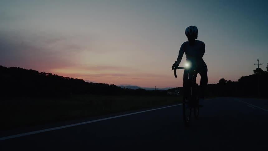 Slow motion silhouette of a female cyclist riding on a rural road against a beautiful sunset sky