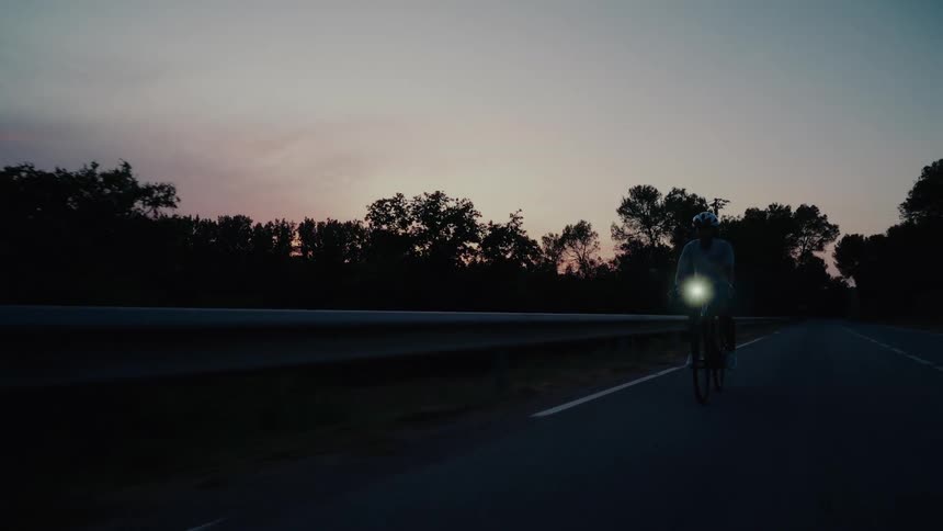 A cyclist with a bright headlight on rides down a deserted country road at dusk