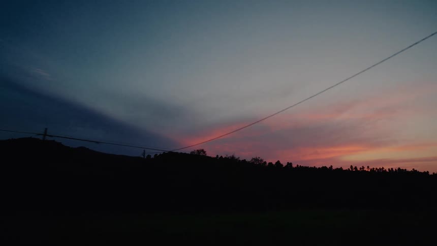 A colorful sunset with a long contrail paints the sky over a dark silhouetted landscape with power lines