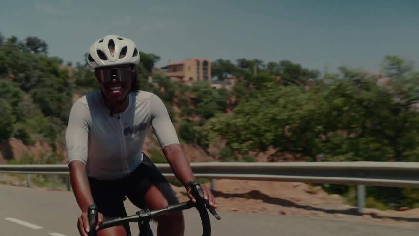 A happy and fit female road cyclist smiles at the camera while riding up a winding mountain road on a sunny day
