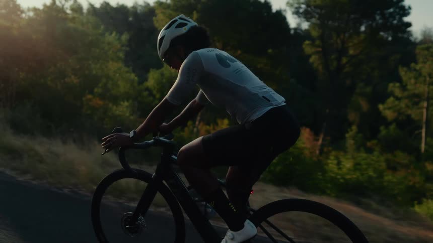A female cyclist in professional gear rides up a sun-drenched country road in slow motion during the golden hour