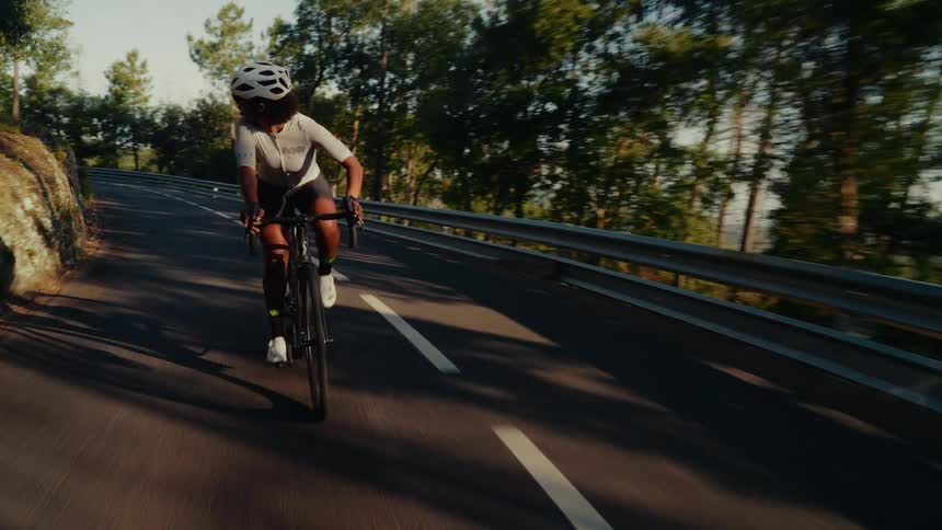 An athletic woman wearing a helmet and cycling gear rides her bike up a paved, winding road through a sunny forest
