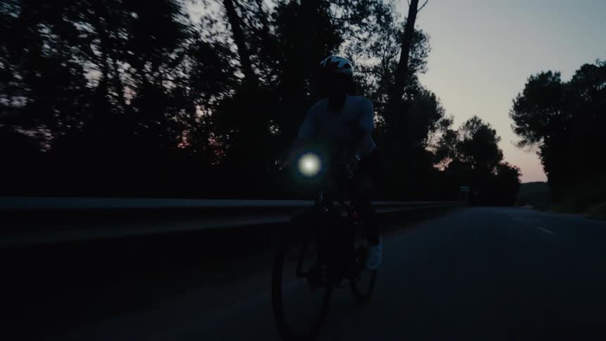 Slow motion shot of a cyclist with a headlight riding a bike on a dark country road at twilight