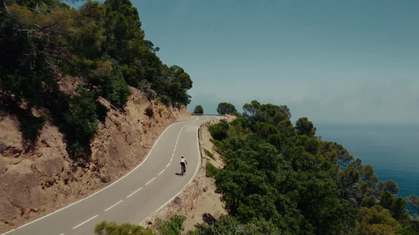 An aerial view captures a lone cyclist riding along a scenic, winding coastal highway carved into a cliffside overlooking the vast ocean