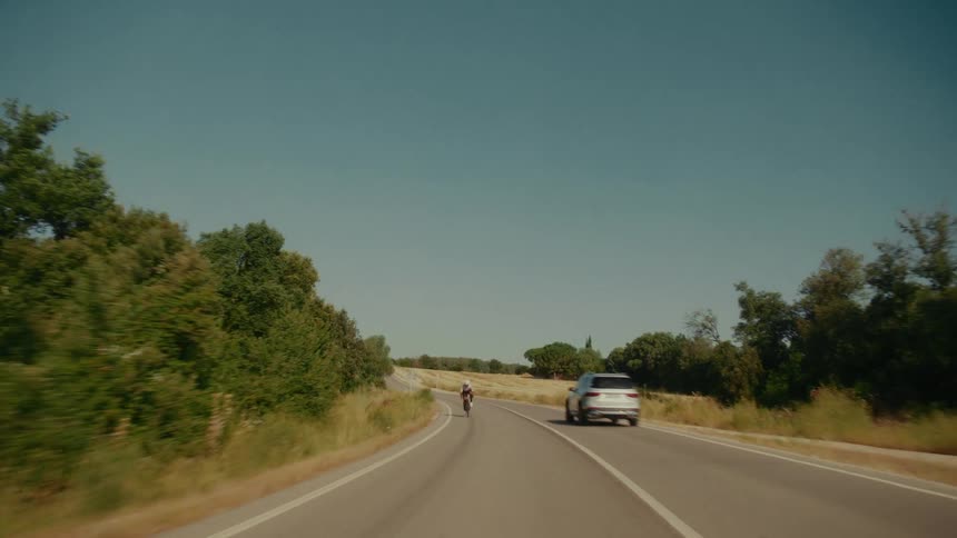 A POV shot from a moving car follows a cyclist riding along a winding country road on a sunny day