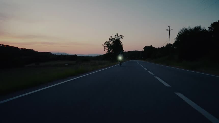 A lone cyclist with a bright headlight rides down a quiet rural road during the twilight hours