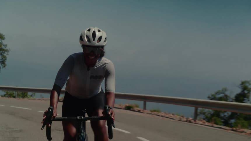 A smiling female road cyclist enjoys her ride up a beautiful mountain pass with the sea in the background