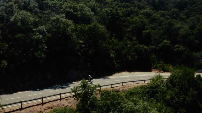 Epic wide shot of a lone cyclist pedaling along a remote road cut into a forested hillside