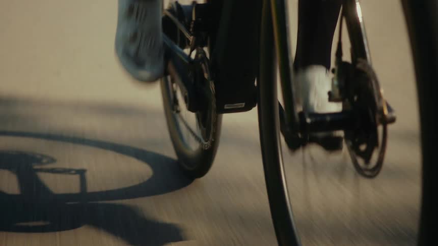 Dynamic close-up of a cyclist's feet in motion, pedaling a road bike as the wheels spin and cast shadows on the road
