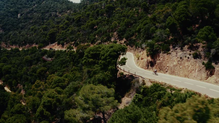 Aerial view of a lone cyclist riding on a winding mountain road surrounded by a lush green forest