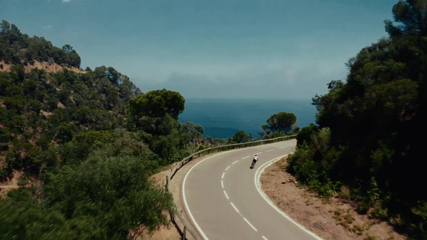 Aerial drone view of a man longboarding down a winding coastal road through a green forest with the ocean in the background