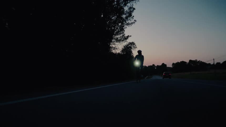 A silhouetted cyclist with a bright headlight rides on a dark country road at dusk as a car passes