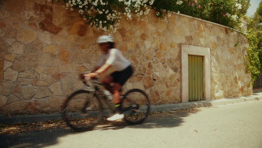 A female cyclist trains on a sunny road, passing by a rustic stone wall adorned with white flowers