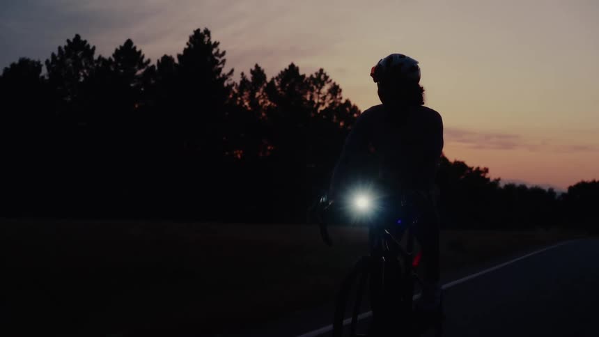 A silhouetted cyclist with a bright headlight rides their bike down a country road during a peaceful sunset