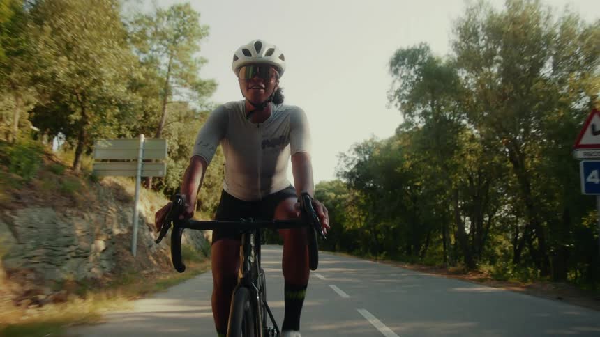 An athletic woman in full cycling gear confidently rides her road bike down a sun-drenched country road
