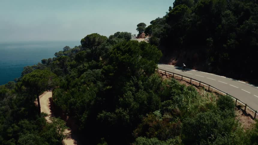 Aerial shot follows a single cyclist riding on a winding road through a lush green forest overlooking the vast ocean
