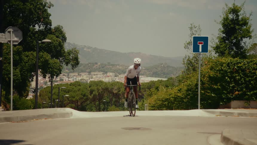 A female cyclist rides her bicycle up a steep hill in slow motion with a scenic town and mountains in the background