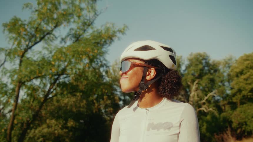 A female cyclist in a helmet and sunglasses smiles happily while surrounded by trees on a sunny day