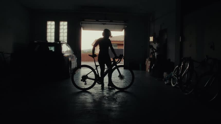 Dramatic silhouette of a cyclist standing with her bike in a garage, illuminated by the open door