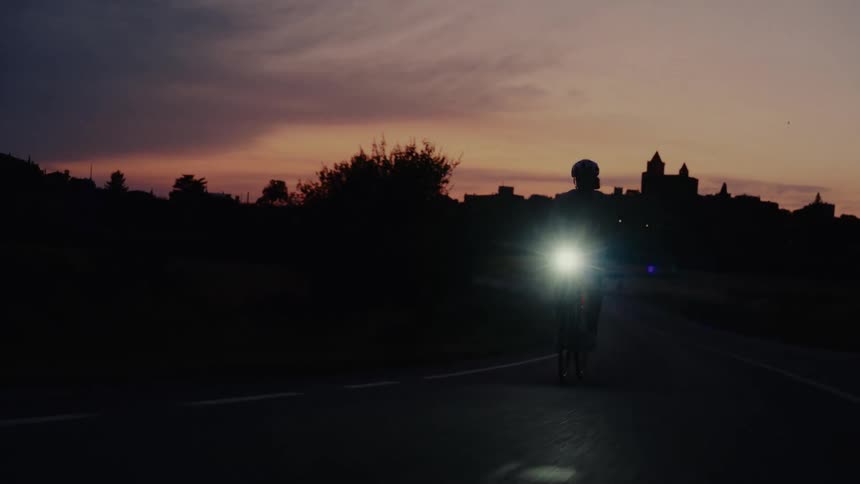 A cyclist with a bright headlight rides on a dark country road, silhouetted against a beautiful sunset sky and a distant village